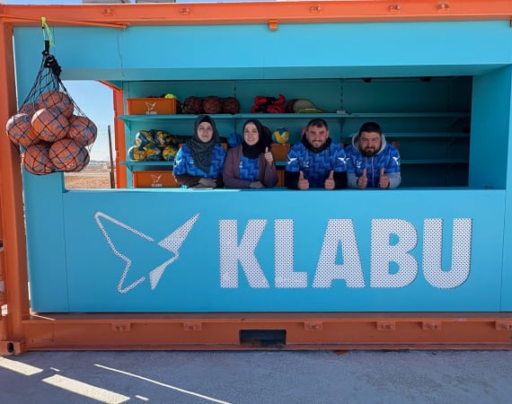 Four people wearing blue shirts stand behind the counter of a bright blue and orange KLABU sports equipment kiosk, with balls and gear visible inside.