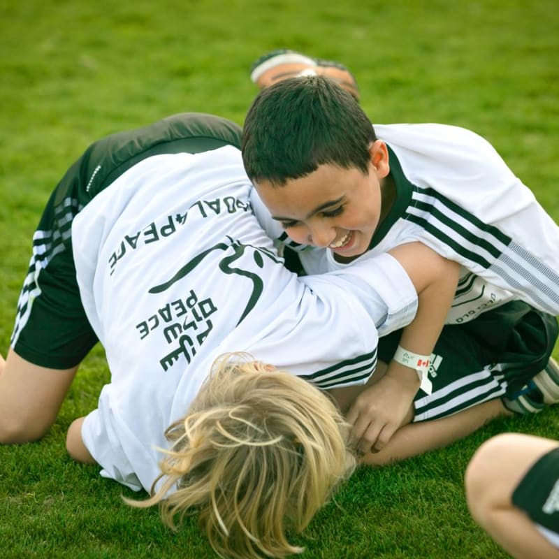 Two children in soccer uniforms laugh and playfully wrestle on the grass during a game, enjoying themselves and smiling brightly.