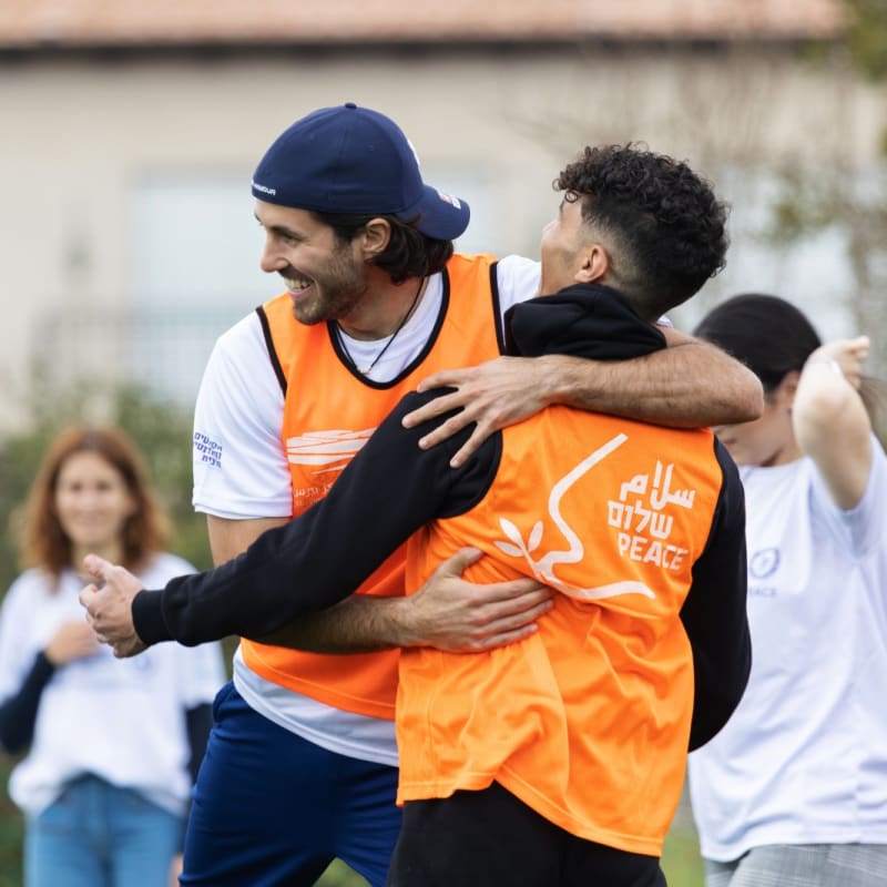 Two young men wearing orange vests, one with PEACE written in English, Hebrew, and Arabic, happily hug during an outdoor activity, while others in white shirts smile in the background.