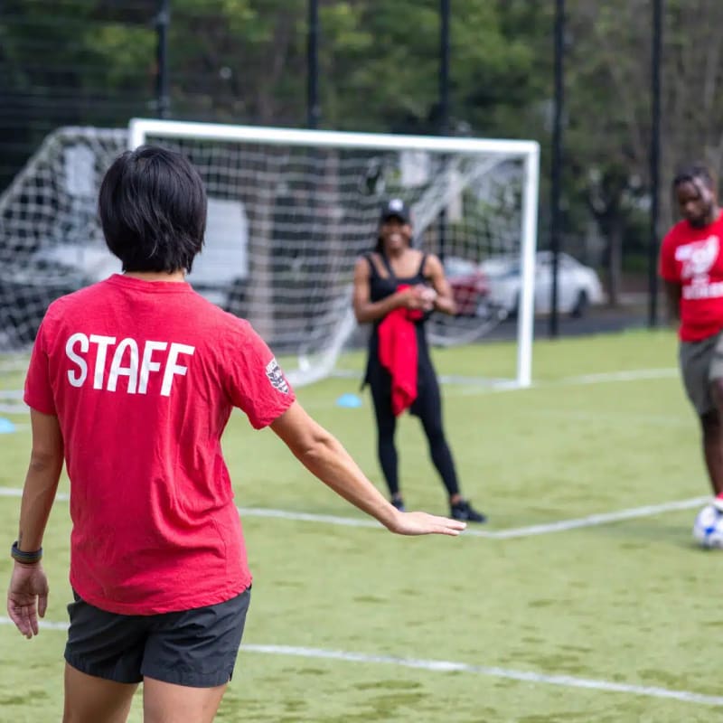 A person in a red shirt with the text "STAFF" on their back oversees a casual soccer game on a grassy field. Players in red and black attire smile and interact near a goalpost.
