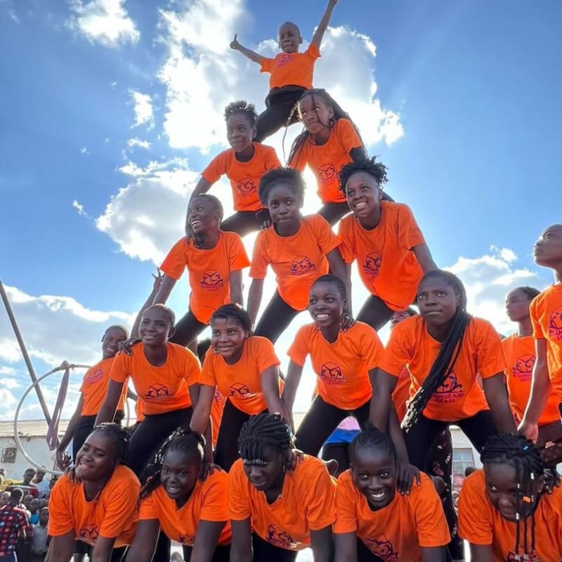 A group of children in bright orange T-shirts form a human pyramid under a sunny blue sky.