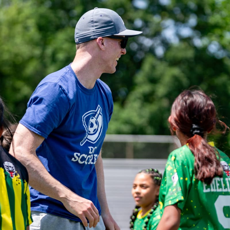 A coach in a blue shirt and cap encourages young soccer players in vibrant yellow and green jerseys on a sunny day.