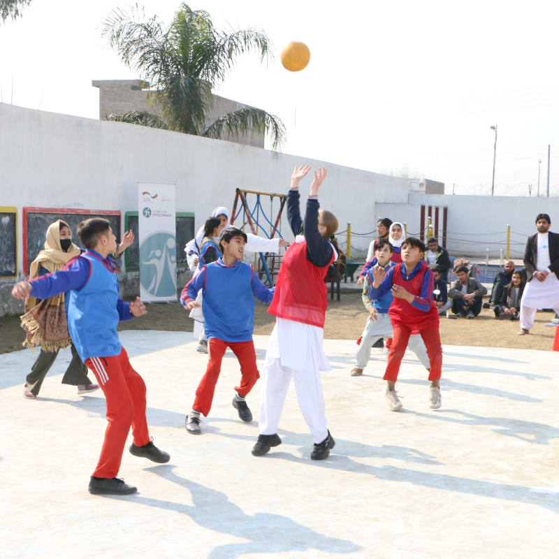 A group of boys and girls, wearing red and blue sports uniforms, play a ball game on an outdoor court while several adults watch from the sidelines on a sunny day.