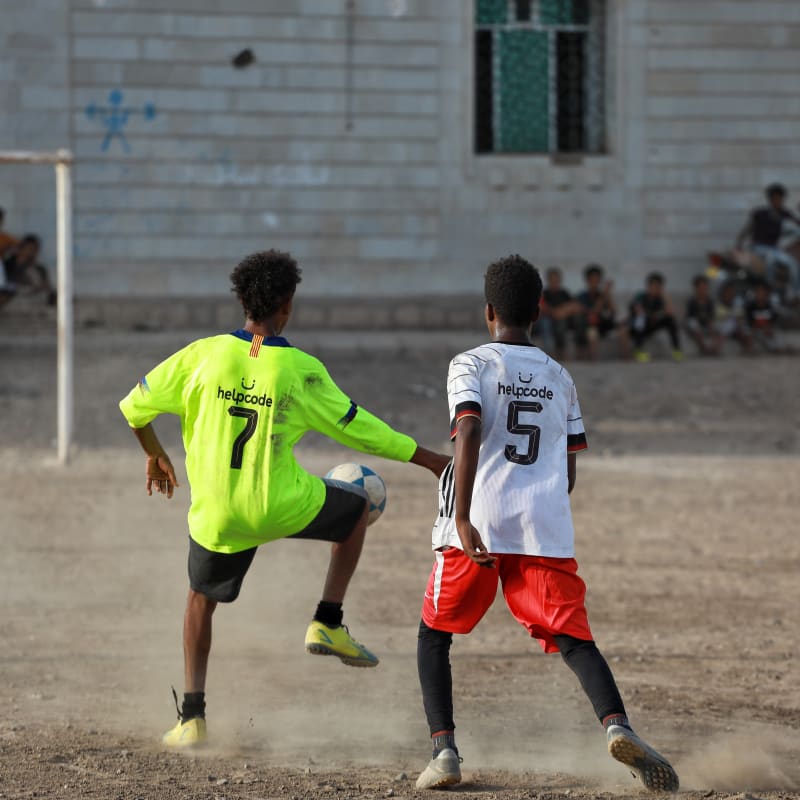 Two young soccer players on a dusty field, one in a neon jersey stopping the ball; others watch from the building in the background.