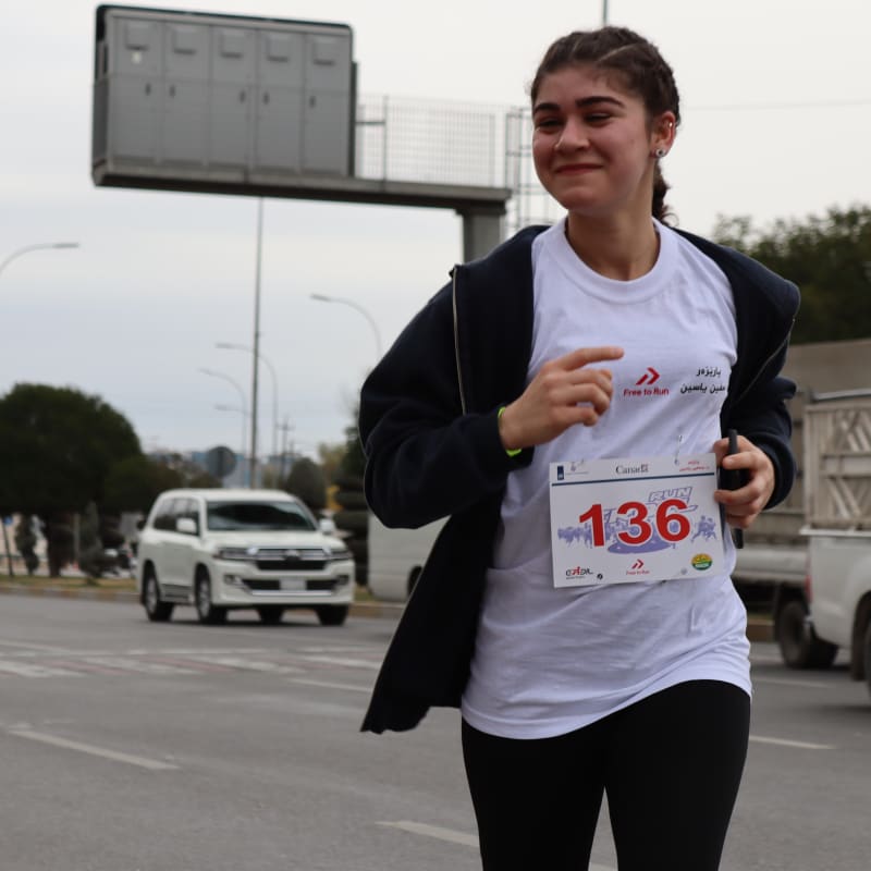 Young woman running a marathon, wearing a white shirt with a race bib number 136, and a black hoodie. She is smiling.
