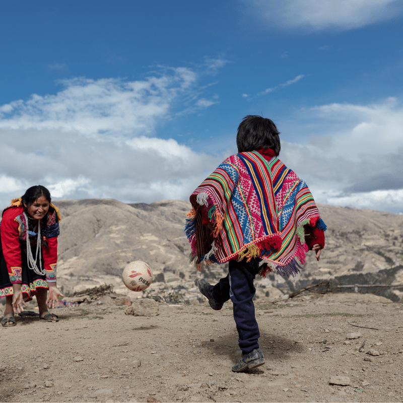 A child wearing a colorful Andean poncho kicks a soccer ball toward a smiling woman in traditional clothing, with mountains and a partly cloudy sky in the background.
