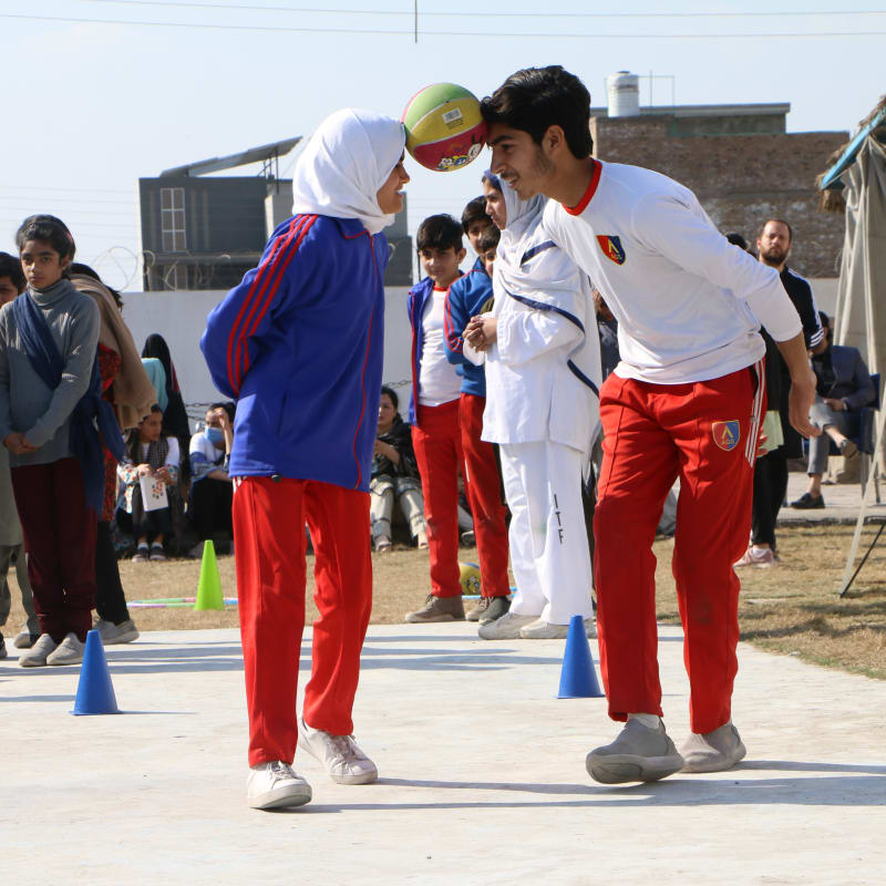 Two children in sports uniforms balance a ball between their heads during an outdoor activity, while other children and adults watch in the background on a sunny day.