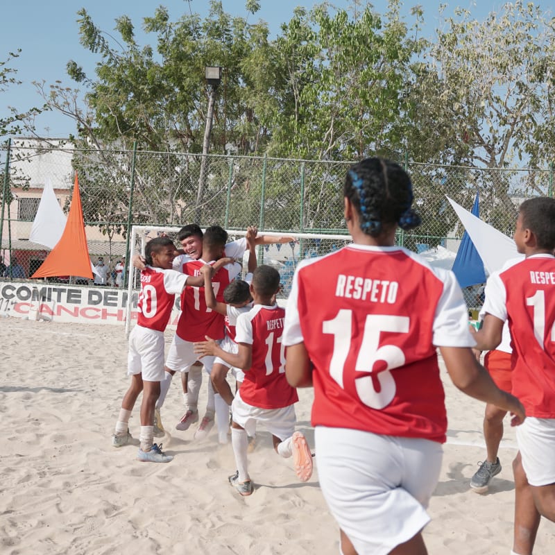 Young soccer players in red and white jerseys celebrate on a sandy field. Banners and trees are visible in the background.