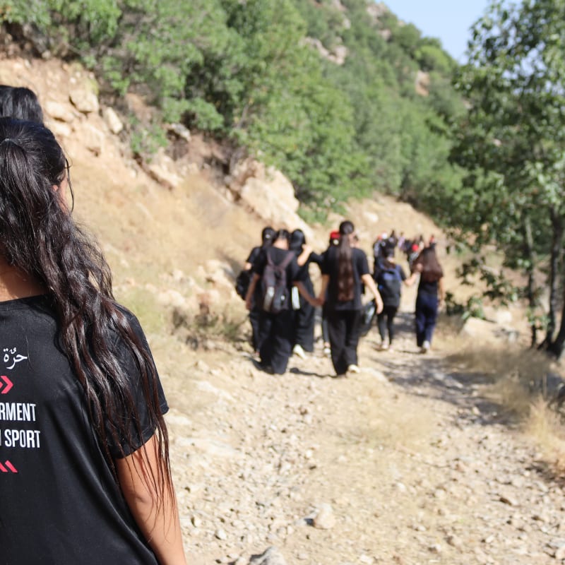 A group of young girls hike along a rocky, tree-lined path in sunlight. A woman in the foreground has "Empowerment Through Sport" text on the back of her black T-shirt.