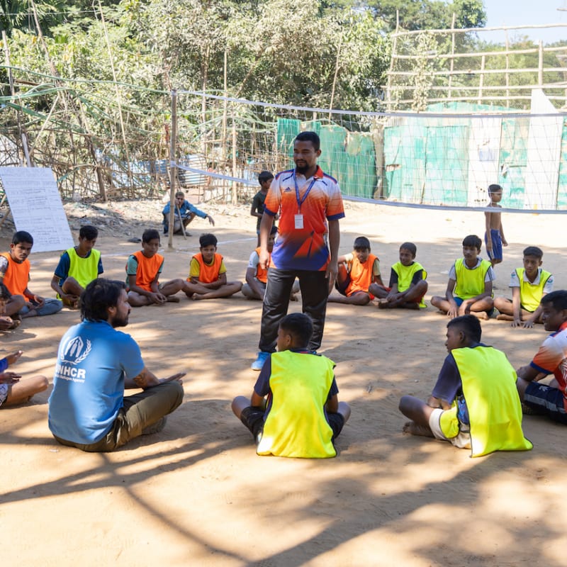 A group of children wearing orange and yellow vests sit in a circle on a sandy ground outdoors, listening to an adult standing in the center. A man in a blue shirt sits among the children. Trees and fencing are in the background.