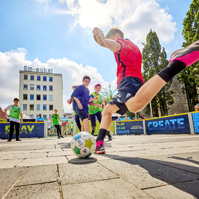 A group of children play soccer outdoors, one in red kicks the ball away from the camera. The sky is bright, with a building in the background.