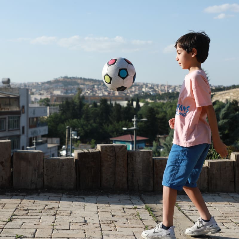 Young boy in a pink shirt and blue shorts plays with a soccer ball outdoors. Cityscape and trees in the background under a clear blue sky.