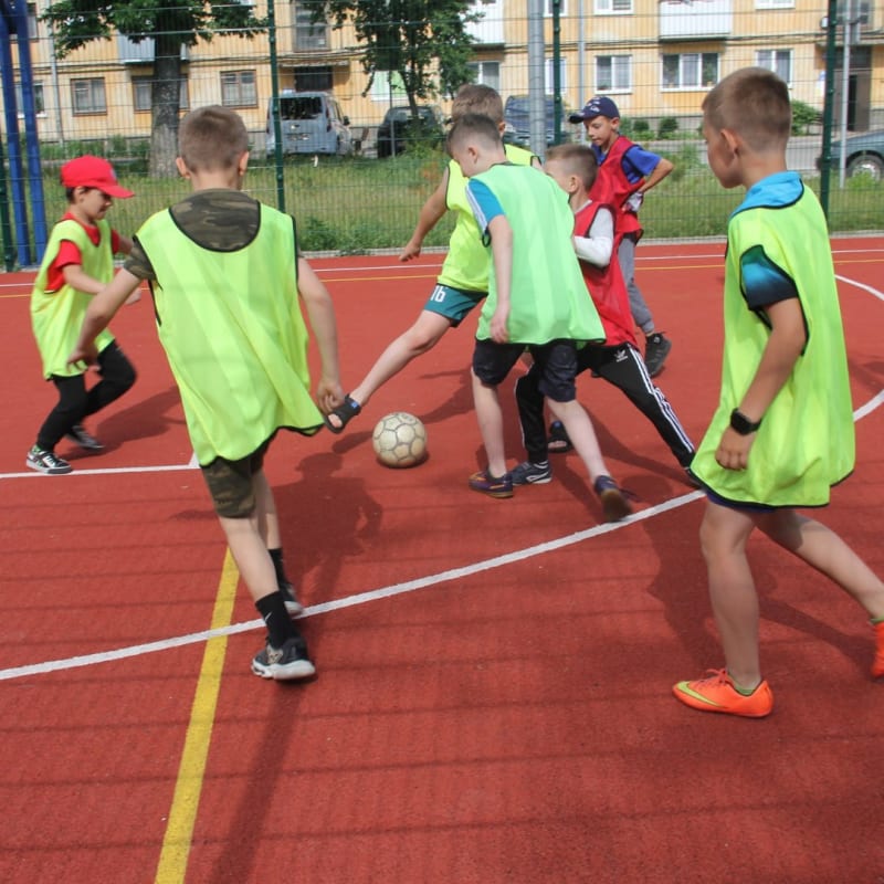 Children play soccer on a red outdoor court, some in neon green vests, one in red.