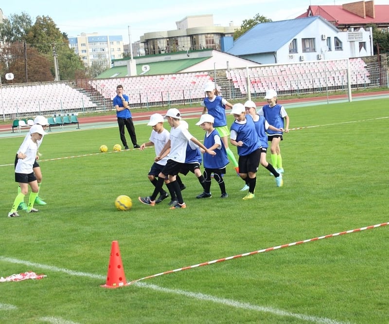 Kids in white and blue jerseys play soccer on a grassy field, surrounded by orange cones. A coach observes from the sidelines.