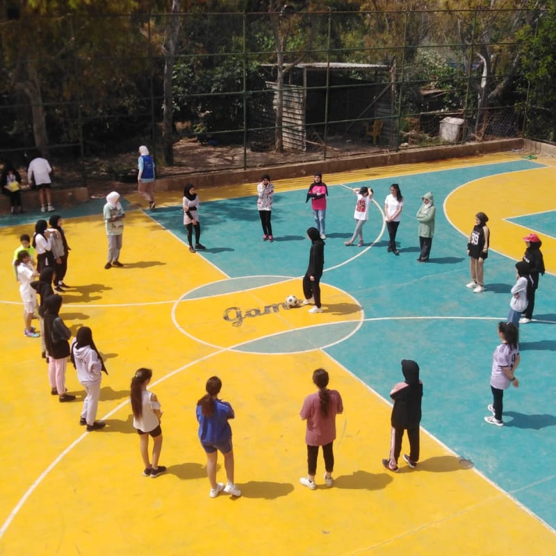 A group of children wearing casual sports clothes stand in a circle on a colorful outdoor court, with a soccer ball in the center.