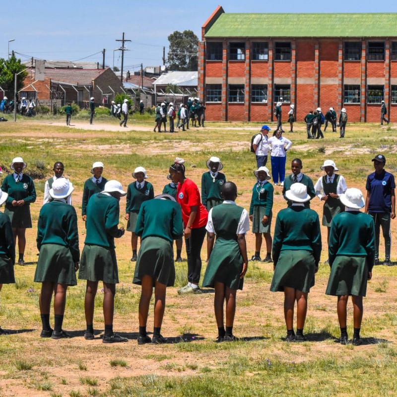A group of students in green uniforms stand in a circle on a grassy field, with others scattered around and a large brick school building in the background. The weather is sunny and bright.