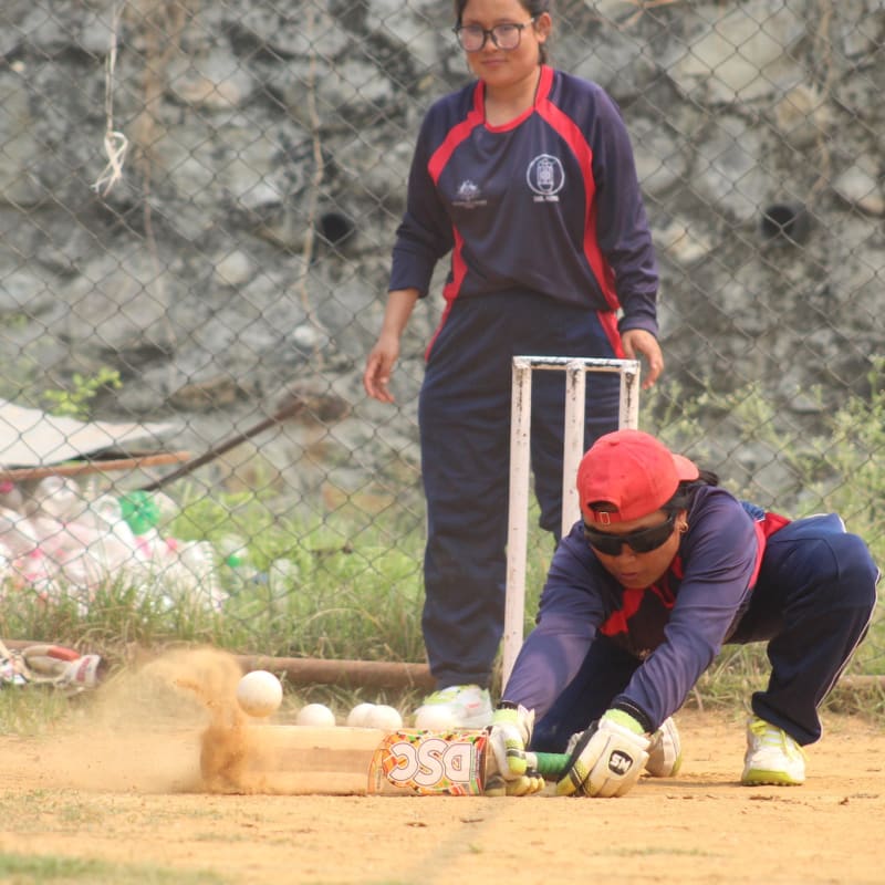 A cricket player in a red cap and sunglasses dives to catch the ball, while another player in matching gear watches. Dust rises from the ground, showing motion.