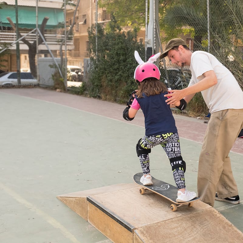 A child wearing a pink helmet with bunny ears skateboards down a small ramp. An adult is assisting her.