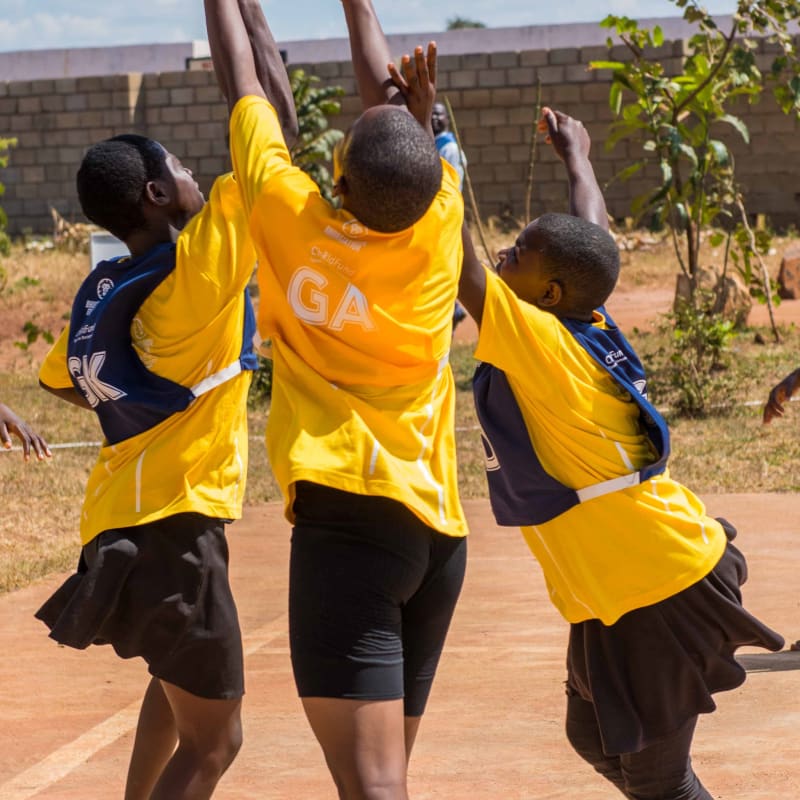 Three girls in yellow and blue sports uniforms jump and reach for a red ball on an outdoor court under a blue sky, with a brick wall and plants visible in the background.