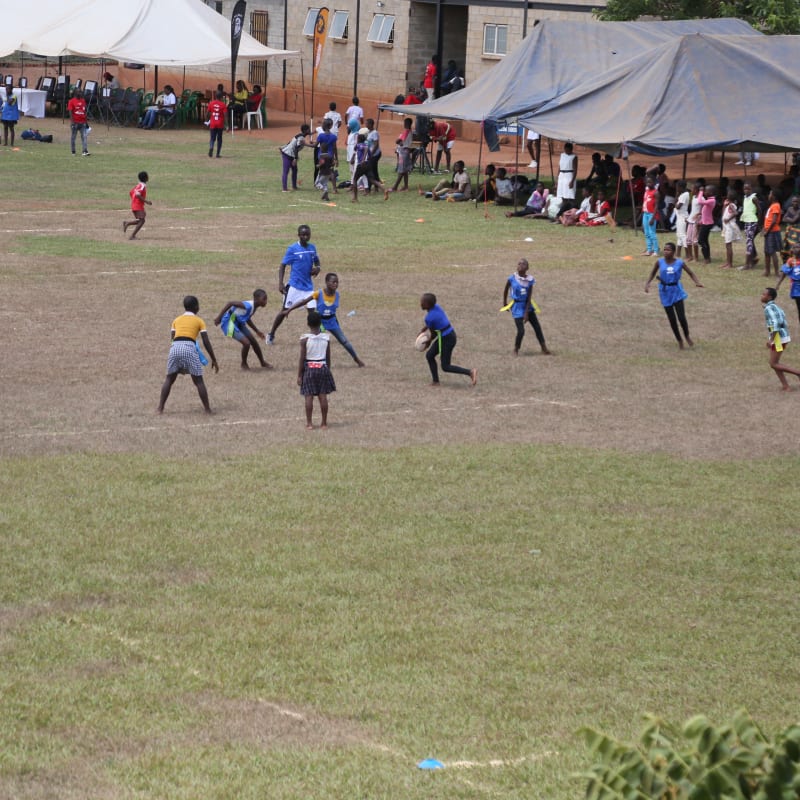 Children play a game on a grassy field, surrounded by people watching under large tents. Some are wearing blue shirts, while others wear different colors. In the background, more people gather near buildings.