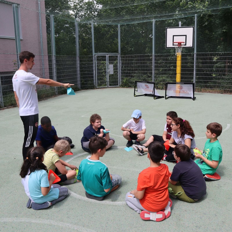 A group of children sit in a circle on an outdoor sports court, listening to an instructor standing and pointing. A basketball hoop and small goals are in the background, with trees and a fence surrounding the court.