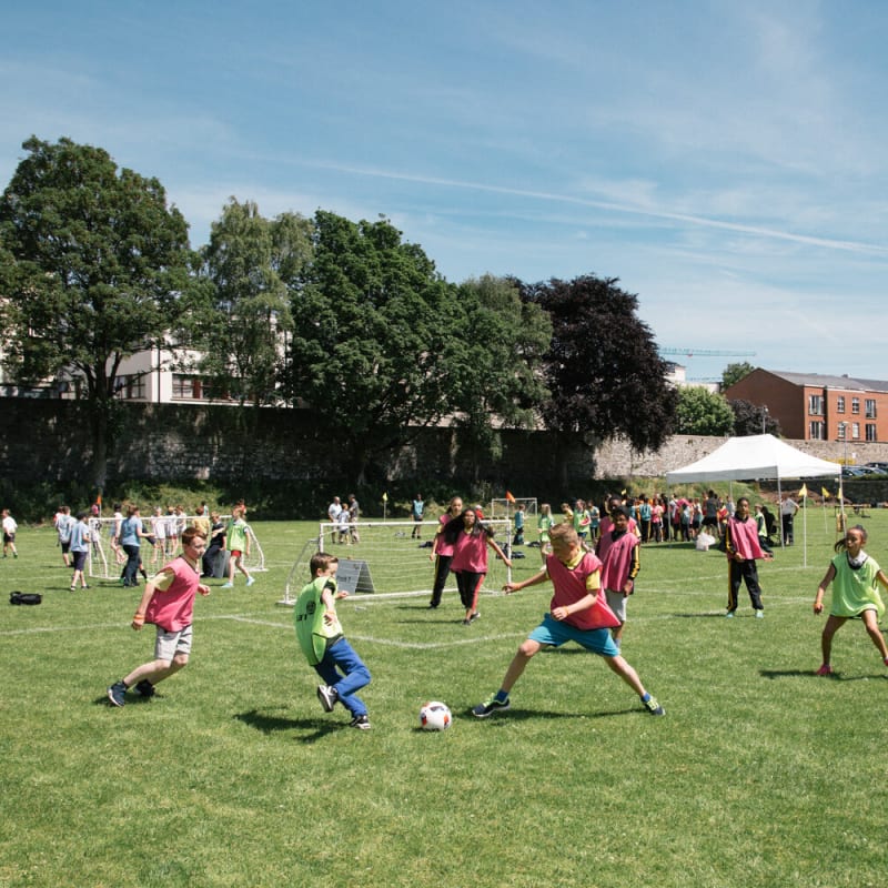 Children playing soccer on a grassy field during a sunny day, with other kids and adults in the background participating in different activities; trees, buildings, and a white tent are visible behind them.