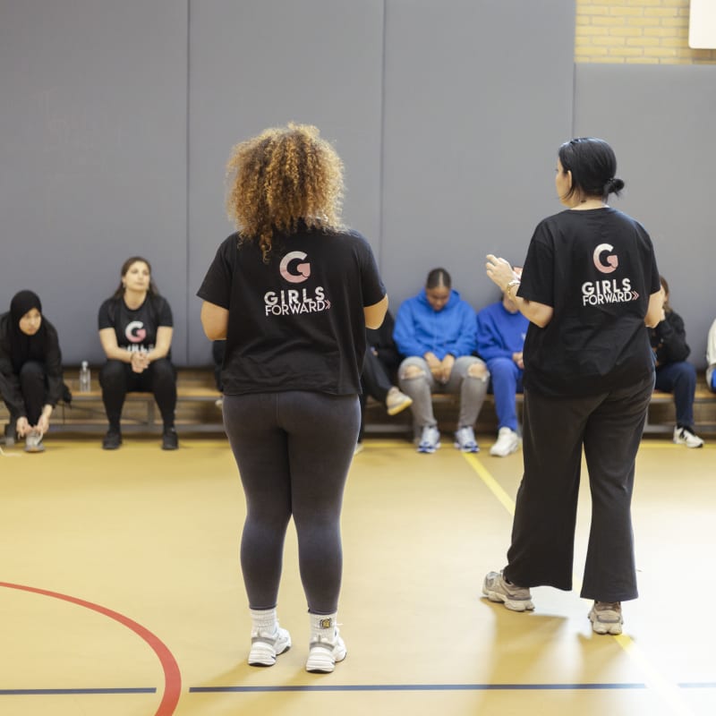 Two women stand in a gym, wearing "Girls Forward" shirts, facing a row of seated individuals.