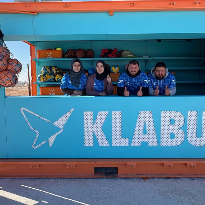Four people wearing blue shirts stand behind the counter of a bright blue and orange KLABU sports equipment kiosk, with balls and gear visible inside.
