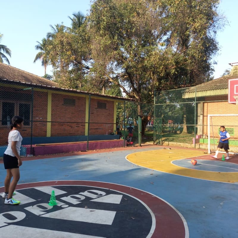 Two girls playing football basketball on an outdoor court surrounded by trees and a wire fence. The court has blue and red markings, and a brick building is visible in the background.