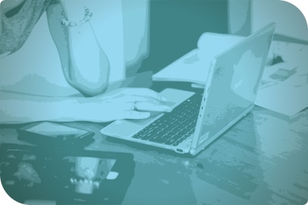 Una mujer está trabajando en su computadora, que está en una mesa con varios dispositivos electrónicos.