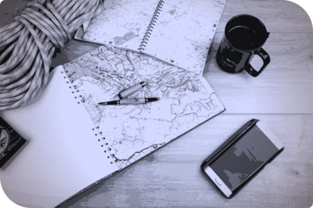 Un libro de mapas y un cuaderno de viajes están en una mesa, junto con un teléfono y una taza.