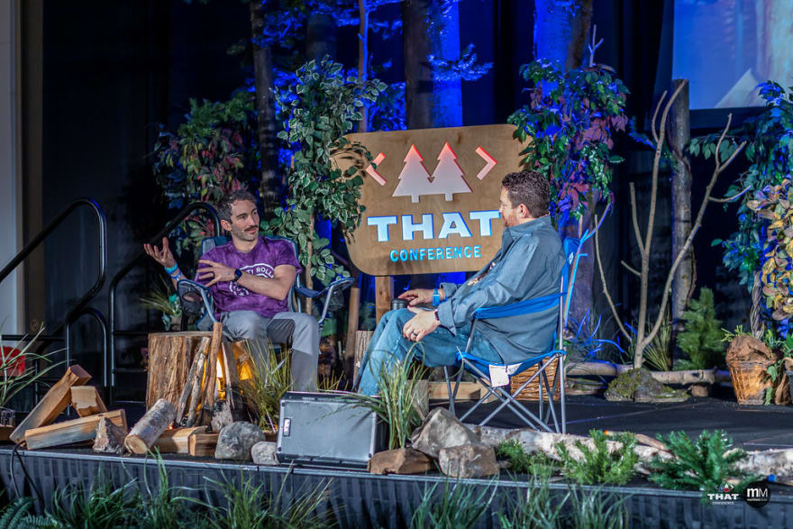 Me sitting with Clark in some camping chairs on the conference stage. We're recording an episode of the podcast and we're surrounded by fake trees, plants, logs, and rocks.