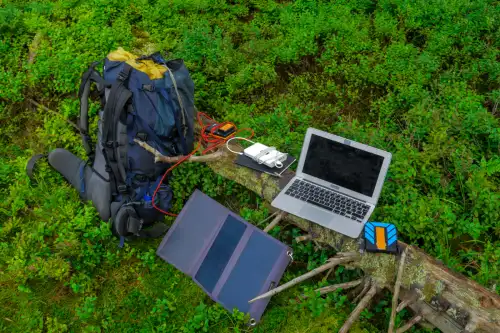 A photo of a backpack leaned against a log on which a laptop, a tablet, and other electronic devices are charging from a portable solar panel.