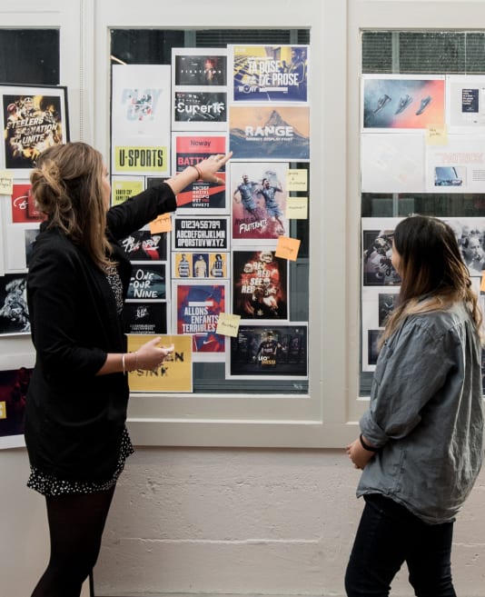 One person pointing at designs stuck on a window with another looking on