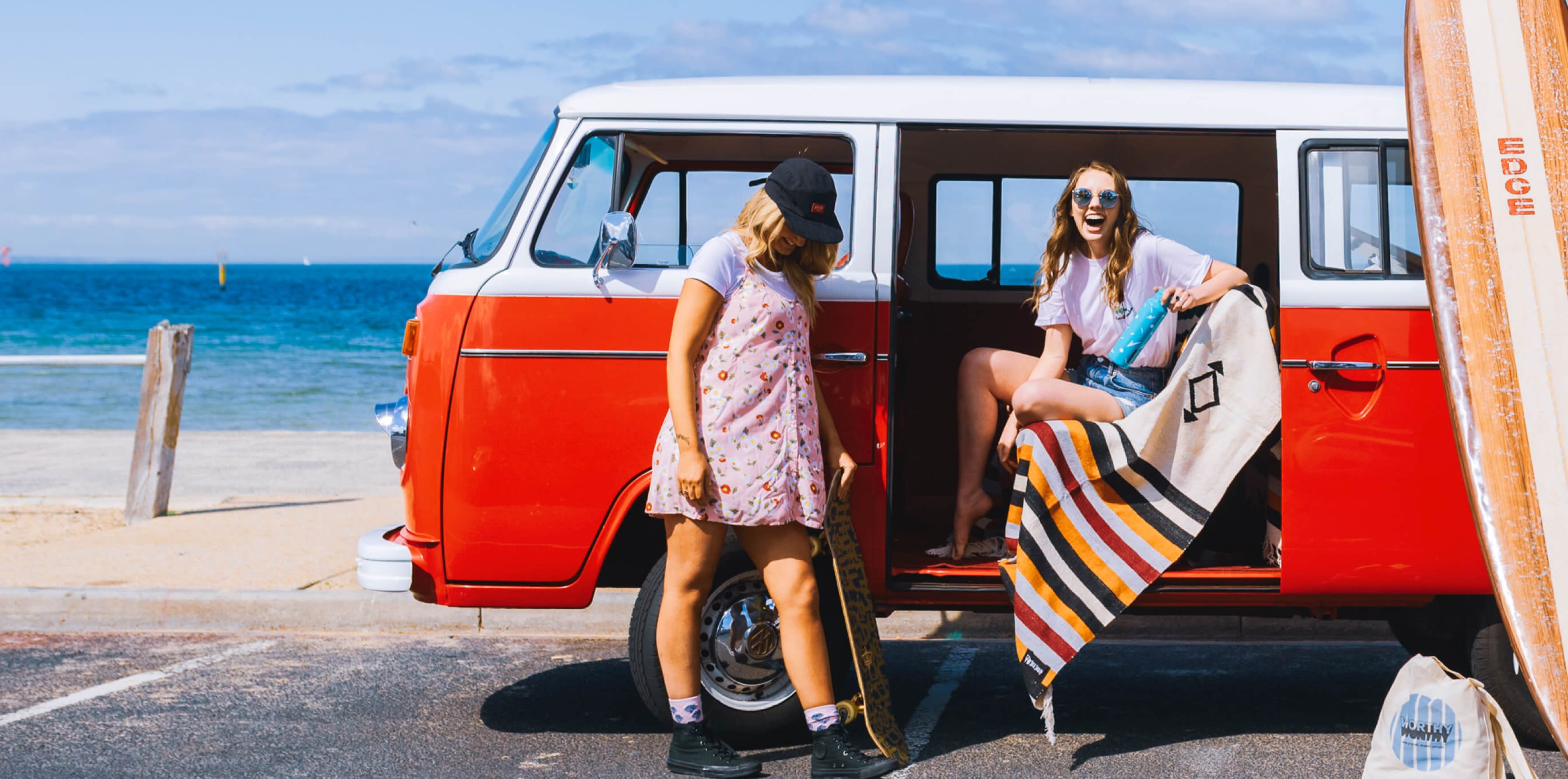Two women standing next to a red VW van, next to the beach.