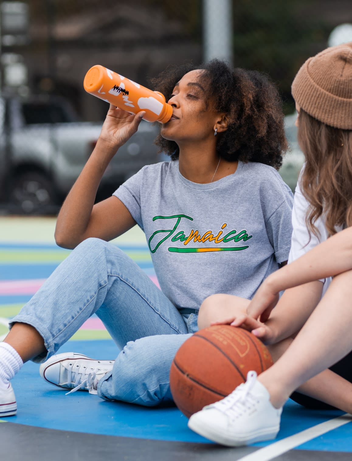 Two people sitting down on a basketball court, one with an orange drink bottle and the other resting as basketball on the court
