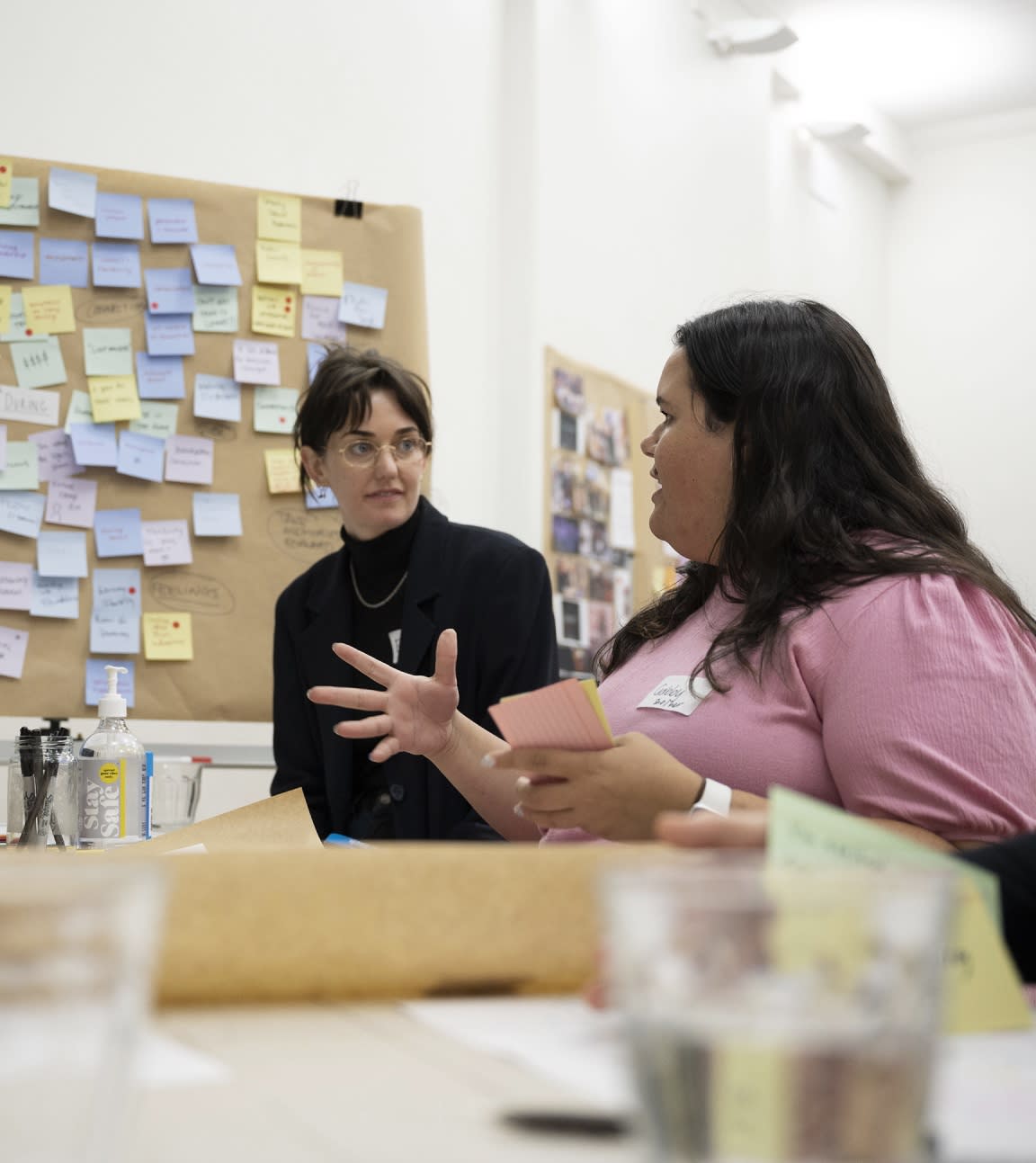 Two people having a discussion at a table with a board of post it notes in the background