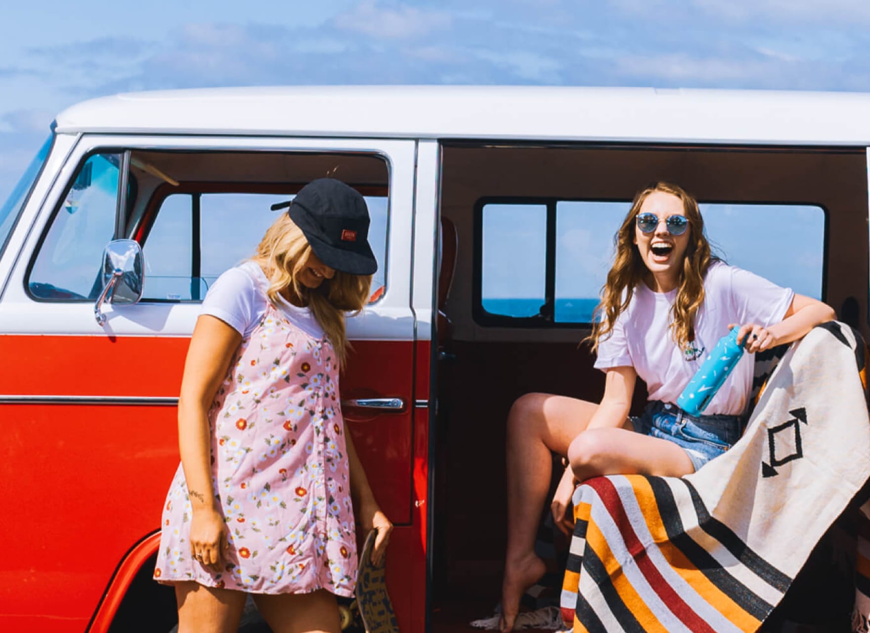 Two women standing next to a red VW van, next to the beach.