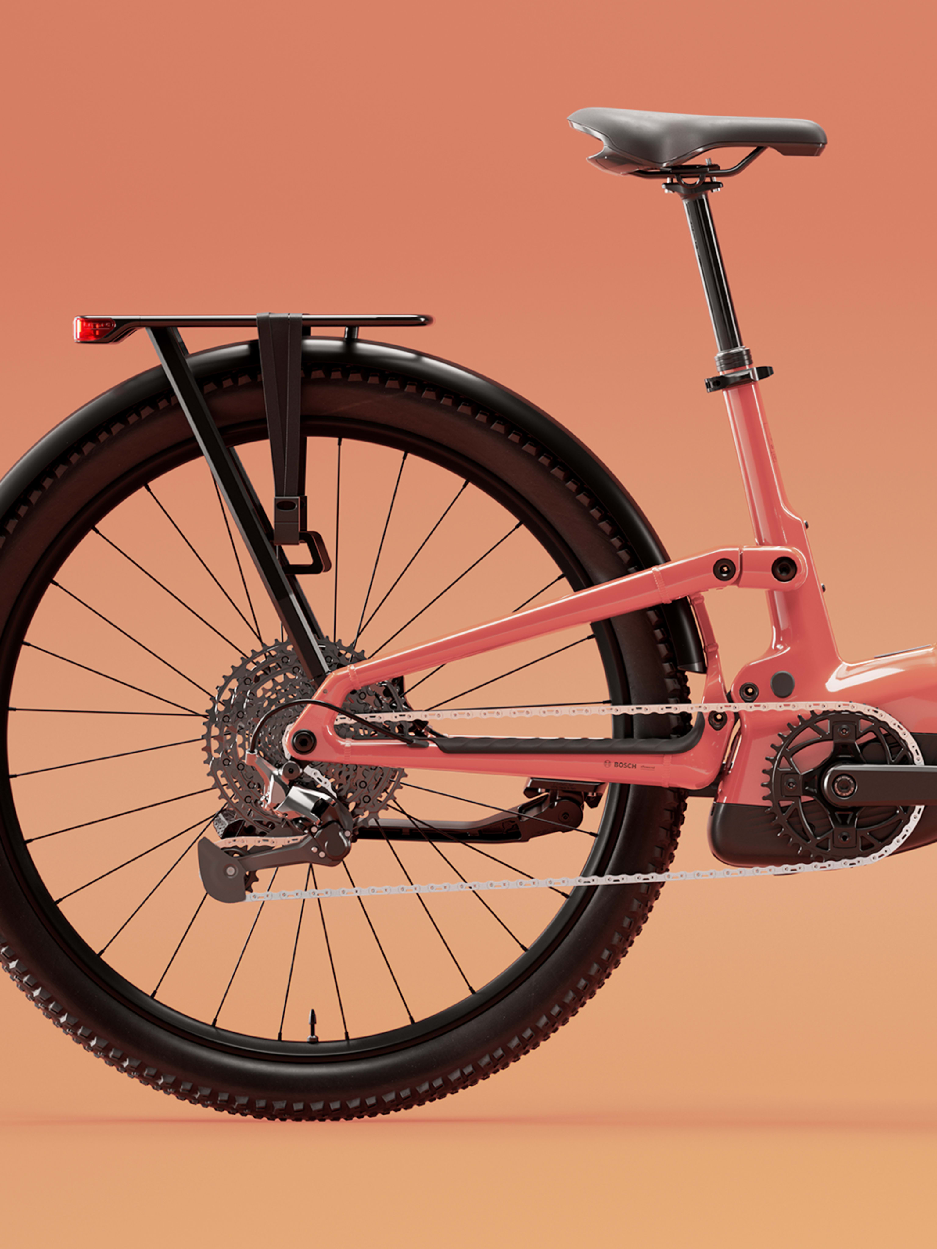 Close-up of a coral-red electric mountain bike drivetrain, showing gears, chain, suspension, and knobby rear tire against a warm backdrop.