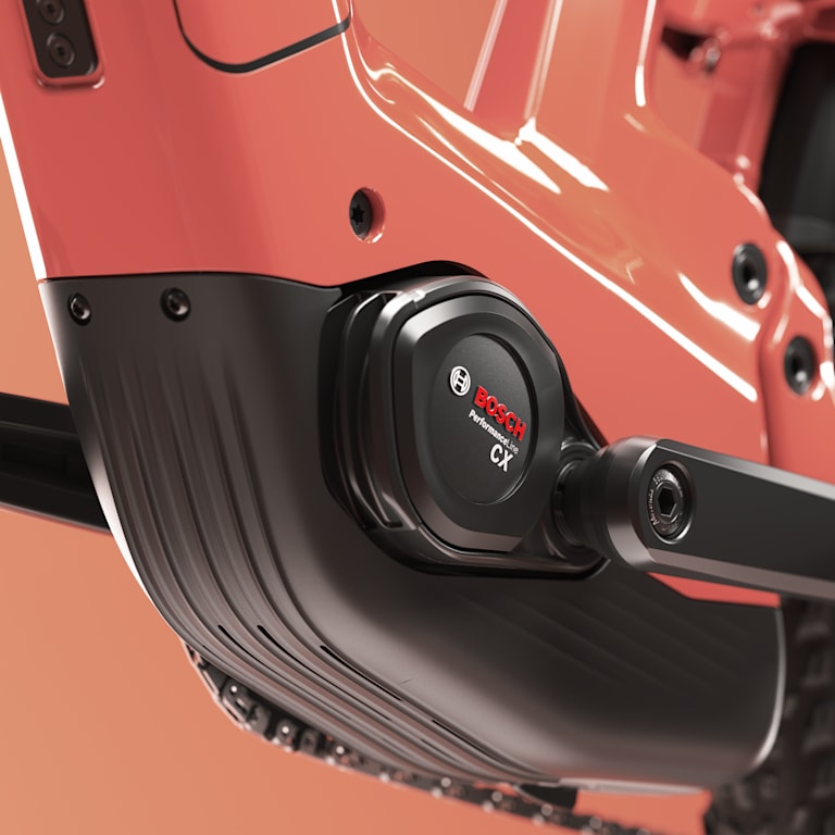 Close-up of a red electric bicycle’s black mid-drive motor, frame, crank area, and rear wheel against a warm peach background; sleek, modern.