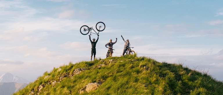 Silhouetted figures celebrate atop a grassy hill with a bicycle raised in triumph, while a vast, cloudy sky stretches over distant mountains.