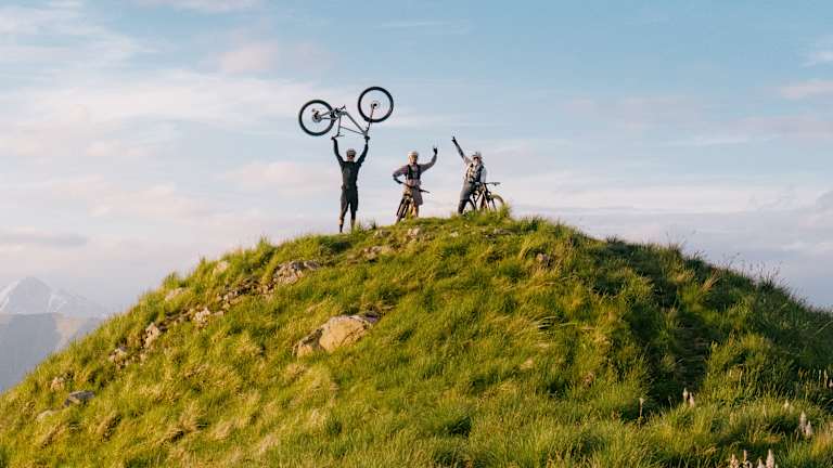 Silhouetted figures celebrate atop a grassy hill with a bicycle raised in triumph, while a vast, cloudy sky stretches over distant mountains.