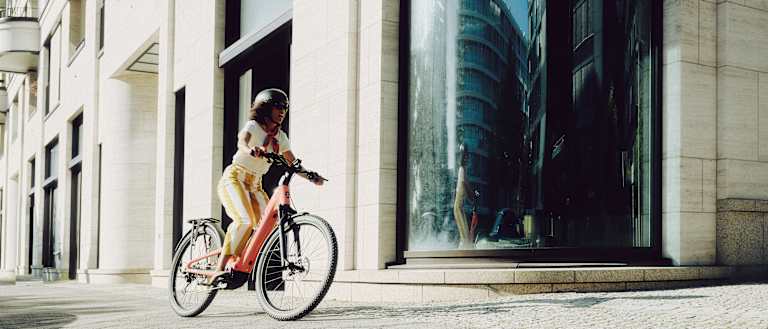 A woman rides a bicycle on a paved sidewalk in front of a modern building with a curved glass window reflecting a city street.