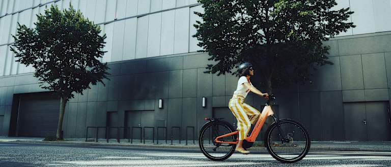 A woman rides an orange electric bicycle past a modern building with trees in the foreground and background.