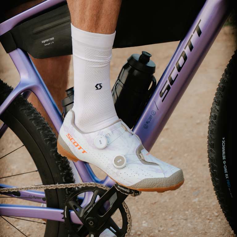 A cyclist's white shoe is clipped into a pedal on a purple SCOTT bicycle, with a dirt path visible in the background.