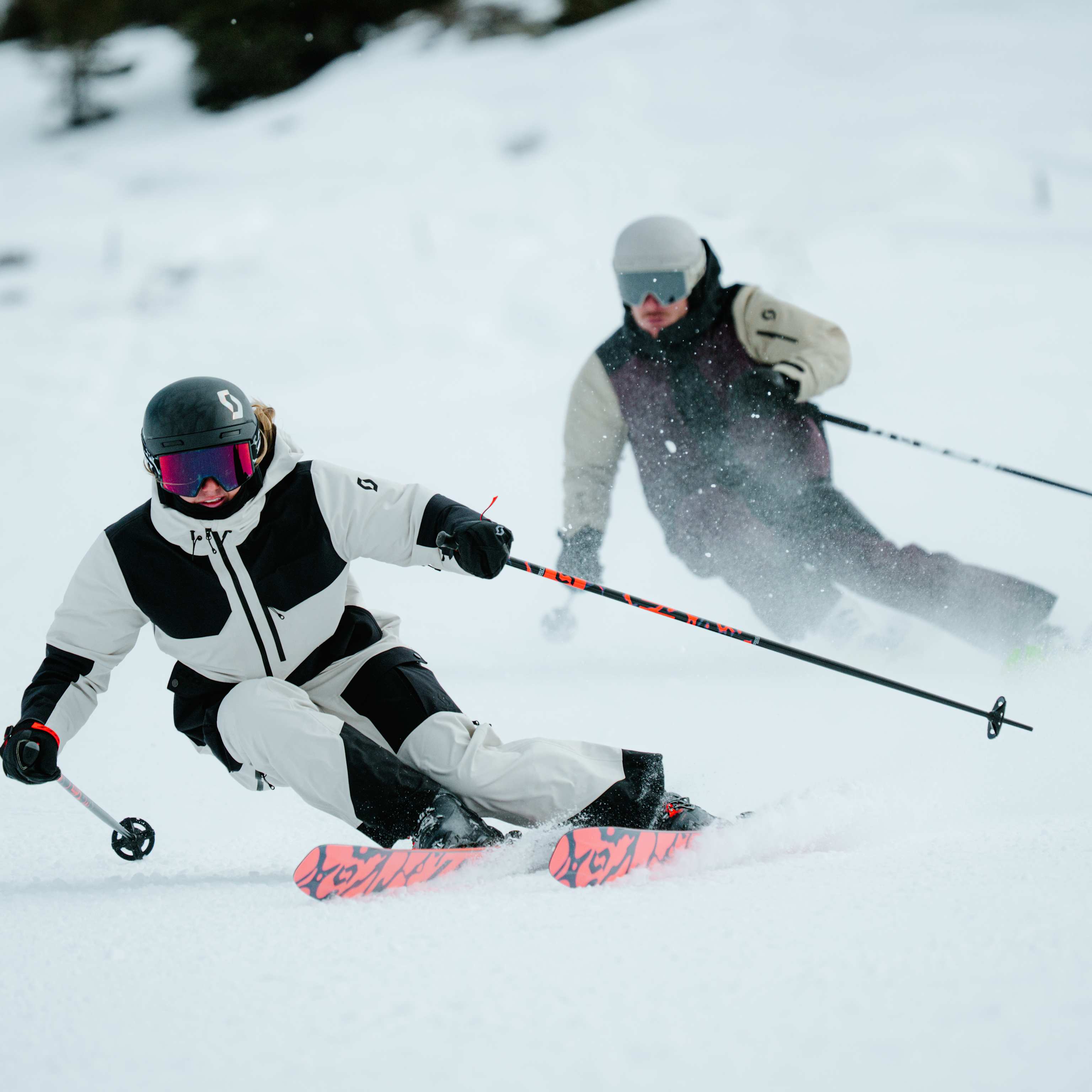 Two skiers carve down a snowy slope, with trees visible in the hazy background.
