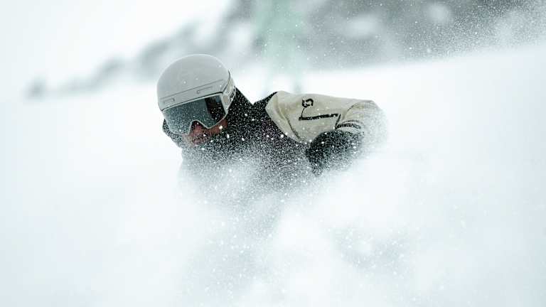 A skier in a white helmet and goggles carves through a cloud of snow, with a blurred snowy background.