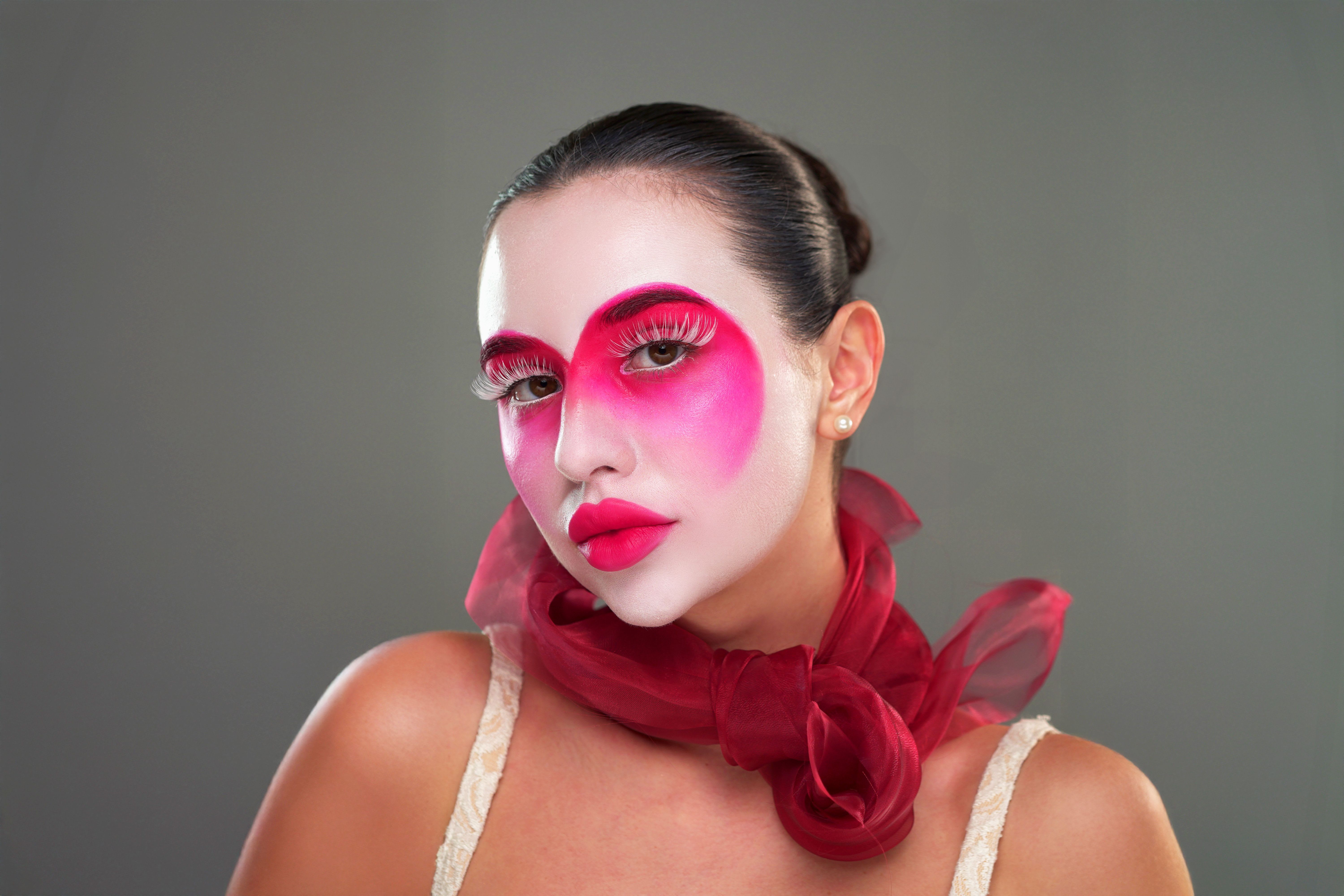Portrait of a woman with bold editorial makeup featuring vivid pink eyeshadow and blush across the eyes and cheeks, paired with defined brows, glossy lips, and a sculptural red tulle accessory around the neck.