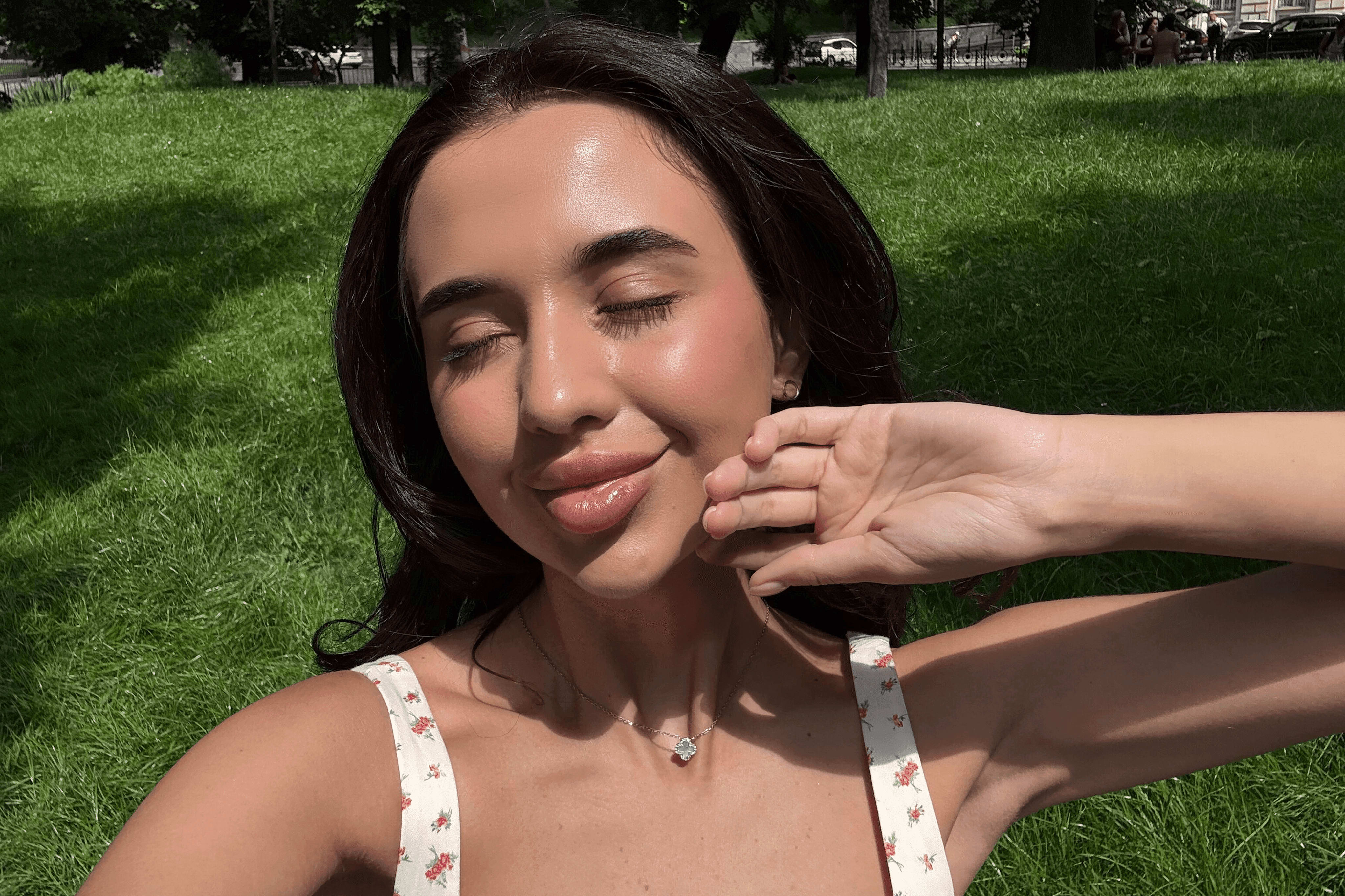 Woman taking a sunlit selfie outdoors in a park, with eyes closed and a relaxed expression, highlighting natural, glowing skin.