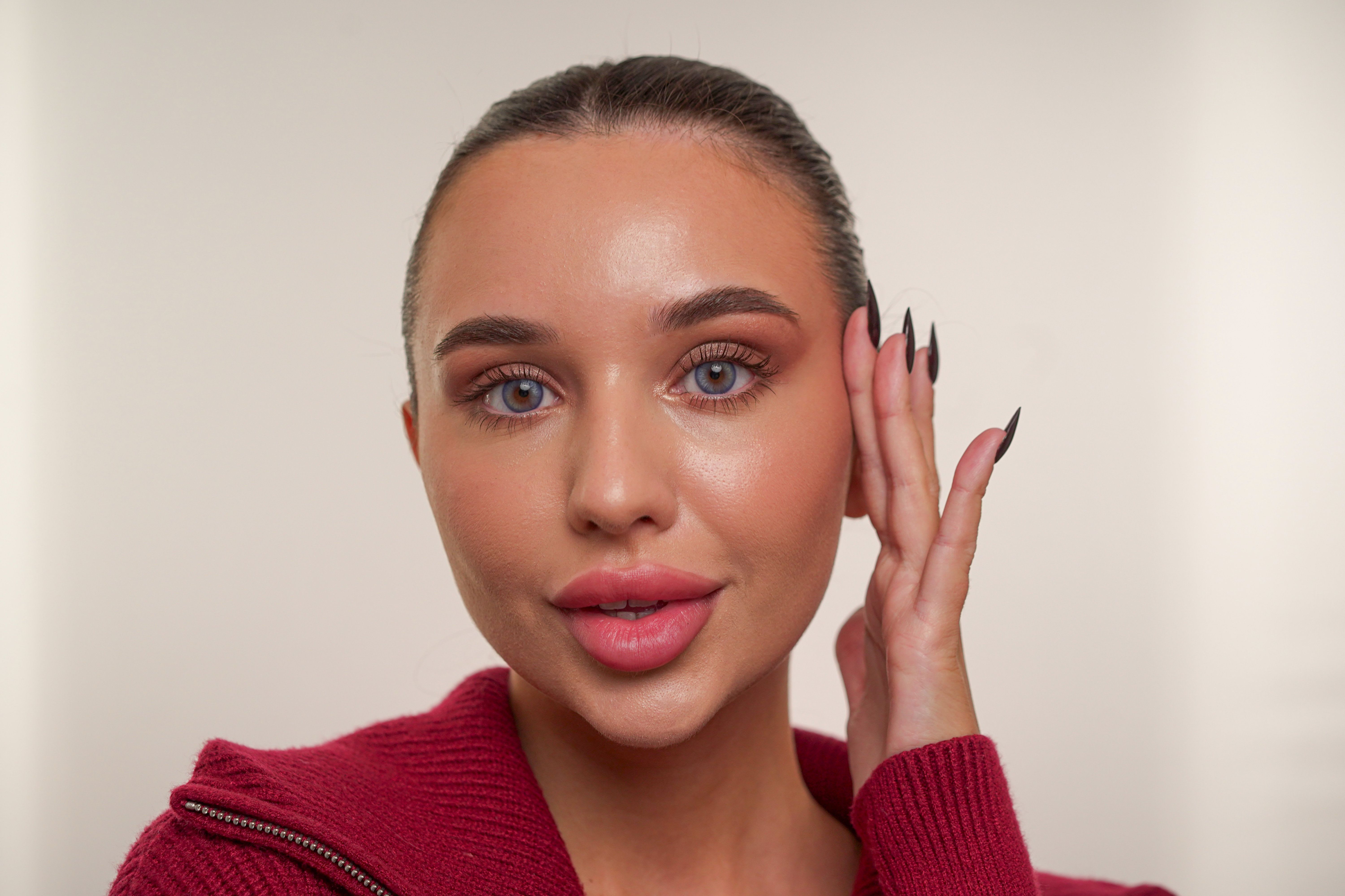 Close-up of a woman with polished glam makeup, featuring defined brows, sculpted skin, and glossy lips, gently touching her face against a neutral background.